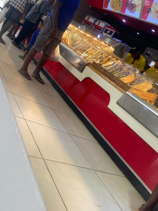 A photograph of customers waiting in line at a bakery or food counter with glass display cases.