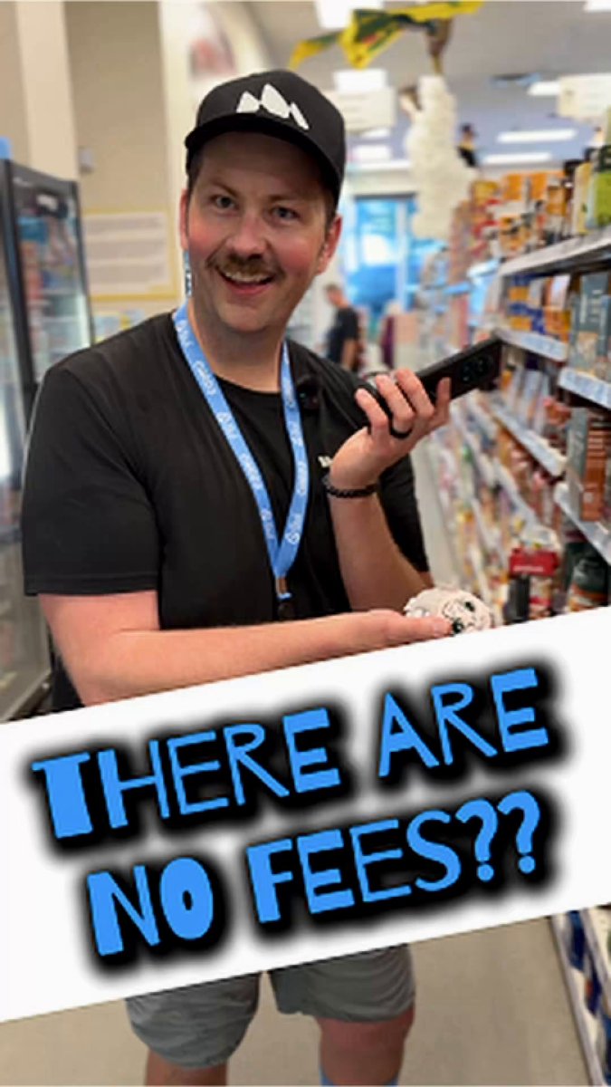 The image features a man standing in front of a grocery store, holding up a sign that reads "There are no fees?". The man has a black shirt and a blue lanyard around his neck. In the background, there are shelves filled with various items such as bottles and cans.