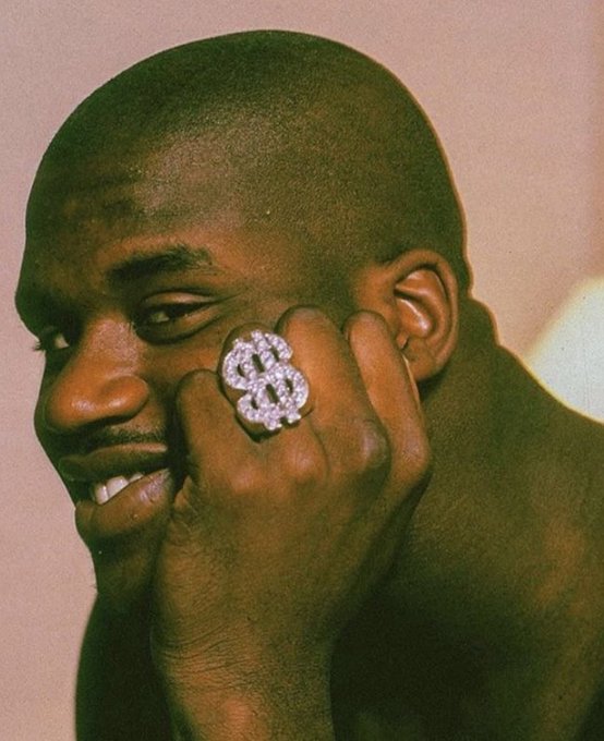 A close-up portrait of a man smiling while resting his face against his hand, prominently displaying a large, ornate dollar sign ($) ring encrusted with jewels.