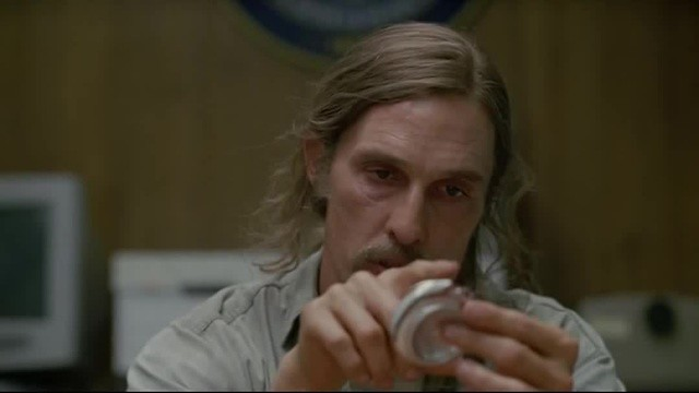 A man with long hair and a mustache holds a small object while looking intently at it, with a seal partially visible on the wooden wall behind him.