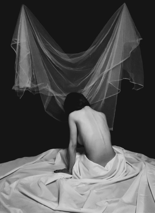 A black and white artistic photograph showing a person sitting with their back to the camera, draped in white cloth, against a dark background with draped fabric above forming a dramatic shape.