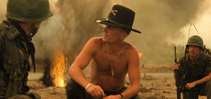 A shirtless soldier wearing a black cowboy hat and a bullet necklace sits calmly amid a war-torn battlefield with explosions in the background, flanked by two fully equipped soldiers in helmets holding rifles. The image's theme of escalating chaos and confidence in the face of danger humorously mirrors the post's commentary on the layered meme-stock-crypto investment chain.