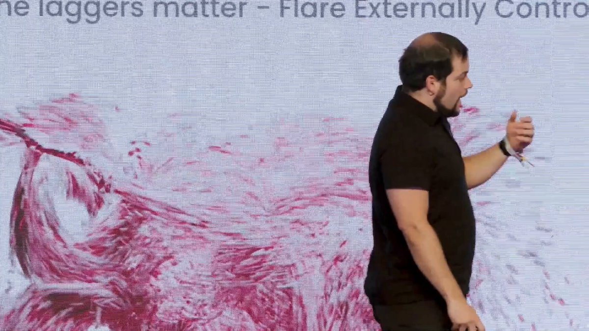 The image shows a man standing next to a large screen with red paint splatters, possibly from a projector or a machine that has been used for an art project. The background of the image features a white wall with black text written on it. The text reads "Flare connects non-smart-contract assets to EVM DeFi while preserving self-custody." This message is related to the man's speech, as he appears to be giving a presentation or lecture about the use of flares in connecting non-smart-contract assets to Ethereum and maintaining self-custody.