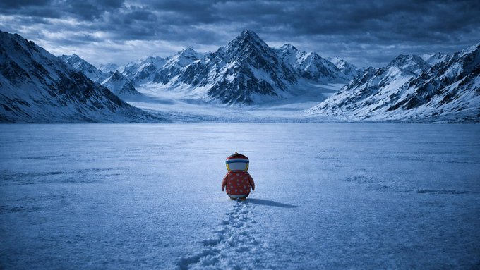 A small character in a red hooded jacket walks across a vast snowy landscape toward towering icy mountains under a cloudy sky.