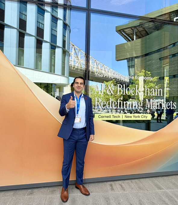 A man in a blue suit gives a thumbs-up in front of a Cornell Tech building window displaying the text 'The Programmable Economy: AI & Blockchain Redefining Markets'.