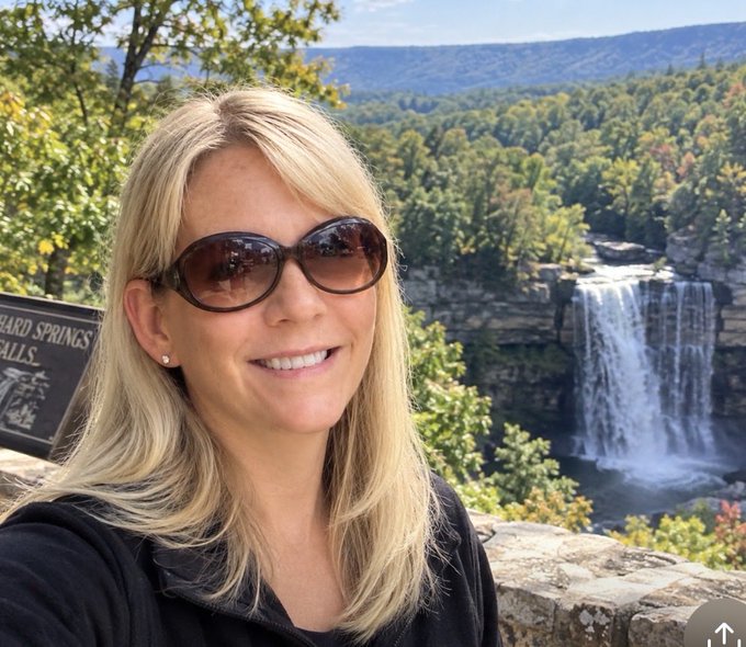 A blonde woman wearing sunglasses poses for a selfie in front of a scenic waterfall and forested mountain landscape, with a partially visible sign reading 'Hard Springs Falls' to the left.
