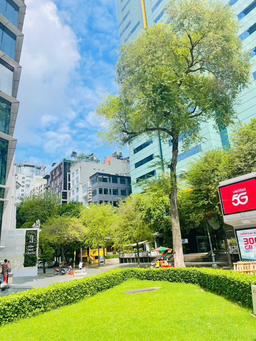 An urban scene featuring modern glass buildings, lush green trees, a manicured lawn, and a Viettel 5G advertisement sign in what appears to be a Vietnamese city.