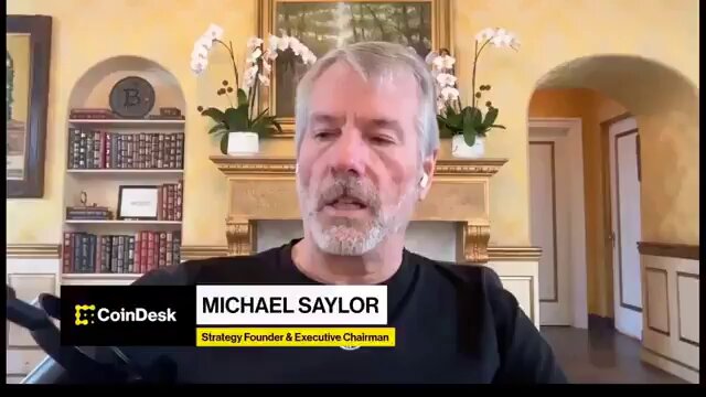 The image features a man sitting down, wearing a black shirt with his name displayed across the front of it. The man appears to be talking about something related to cryptocurrency or blockchain technology. In the background, there are several potted plants placed around the room, and a bookshelf filled with numerous books can also be seen.
