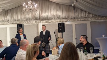 The image shows a group of people gathered around a dining table for a special dinner event. There are at least ten people visible in the scene, engaged in conversation and enjoying each other's company. A man stands up and gives a speech or presentation, capturing everyone's attention. The room is well-lit with chandeliers hanging from the ceiling, creating an elegant atmosphere.

The table is set with various items such as wine glasses, cups, forks, knives, and bowls filled with food. There are also several bottles placed on the table, likely containing drinks for the guests. A chair can be seen in the foreground of the image, indicating that it's a comfortable seating area for the attendees.

The overall setting suggests an intimate gathering where people have come together to share ideas, discuss important matters, and enjoy each other's company over good food and conversation.