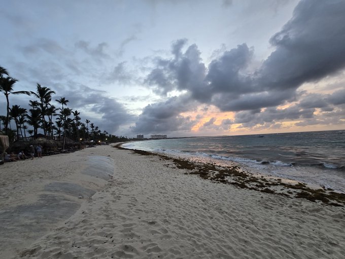 The image shows a beach scene with a cloudy sky above, sand, water, and palm trees. The photo appears to have been taken during sunset or sunrise, as indicated by the warm colors of the sky. The caption "GM X Happy Pre-Friday May today shine bright May today keep you bullish May today keep you smiling" suggests that the beach is likely a popular destination for tourists who want to enjoy the beautiful scenery and take memorable photos.

The image also features several people scattered across the beach, some closer to the water's edge while others are further inland. The presence of these individuals indicates that this location is a popular spot for relaxation and leisure activities during the summer months.