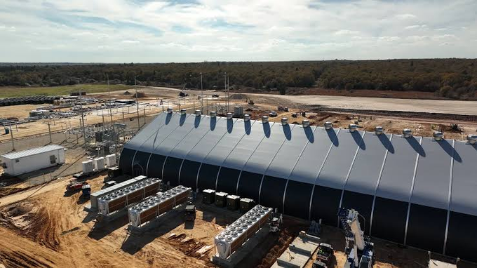 An aerial view shows a large, curved-roof industrial facility under construction in a rural Texas setting, with rows of cooling units and electrical equipment visible alongside the structure, illustrating the type of large-scale data center infrastructure that Anthropic's $9 billion, 20-year lease deal with Riot Platforms aims to repurpose from Bitcoin mining to AI computing.