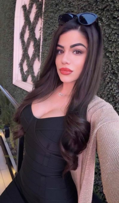 A portrait of a woman with long, wavy dark hair posing in a black top with sunglasses resting on her head, standing in front of a green ivy wall.