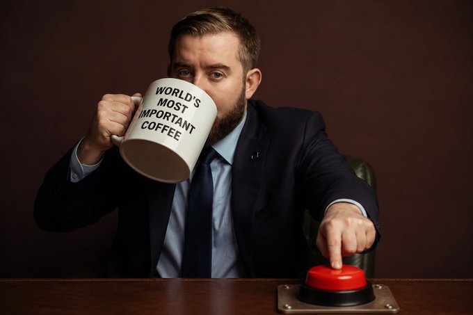 A confident man in a suit casually sips from a mug labeled "WORLD'S MOST IMPORTANT COFFEE" while pressing a large red button, capturing the bold, decisive energy of @SergeyNazarov's call to "start the rerating process now."