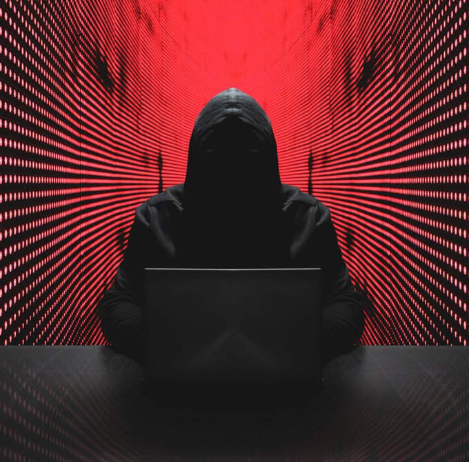 A dark silhouette of a hooded figure sitting at a laptop against a red digital background with radiating light patterns.