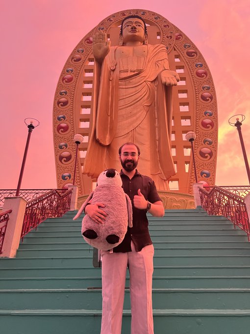 A man with a beard holding a large stuffed sea lion plush toy, giving a thumbs up in front of a large golden Buddha statue at sunset.