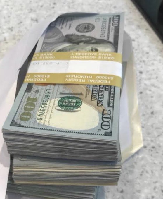 A photo of a large stack of US $100 bills bound with yellow Federal Reserve straps labeled '$10,000 HUNDRED' sitting on a speckled countertop.
