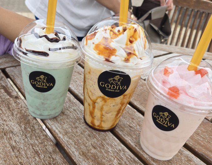 Three Godiva frappes in plastic cups with dome lids and yellow straws rest on a wooden table; the drinks are mint green with chocolate drizzle, caramel with whipped cream, and pink with strawberry pieces.