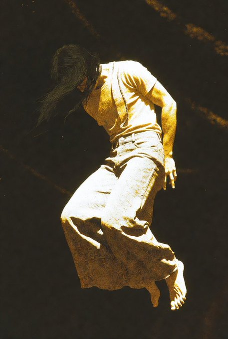 A sepia-toned artistic photograph of a woman with long hair, wearing a t-shirt and skirt, captured in a dramatic downward motion against a dark background.