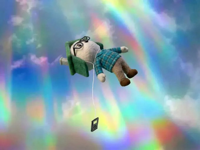 A whimsical 3D illustration of a stuffed doll wearing glasses and a green plaid shirt, appearing to float or fall through a colorful, rainbow-hued cloudy sky. The doll holds a string attached to a small cassette tape dangling below it.