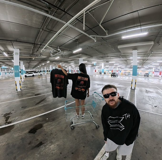 A man in a black hoodie stands in a parking garage while a woman on a shopping cart holds up a black 'METEORA' t-shirt.