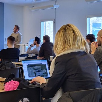 An indoor gathering or class with people seated facing a speaker at the front, while a woman in the foreground works on a laptop displaying code.
