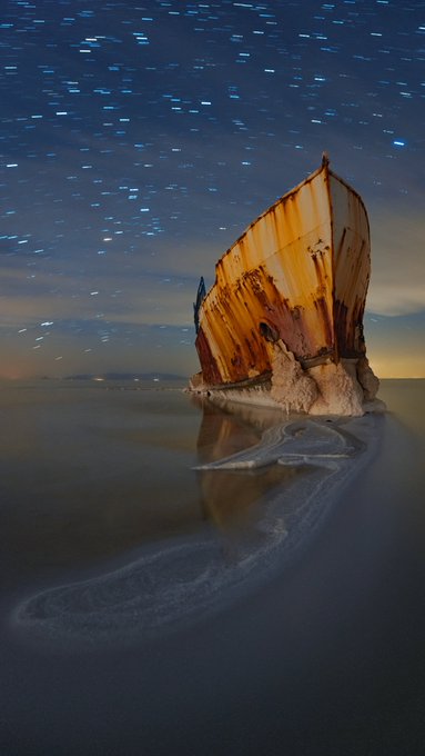 A long-exposure photograph of a rusted shipwreck on a flat salt lake under a starry night sky with streaking star trails.