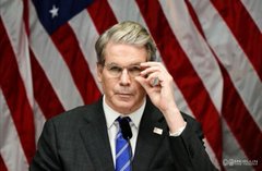 A man in a dark suit adjusts his glasses while standing in front of multiple American flags, appearing to be at a government hearing or press conference.
