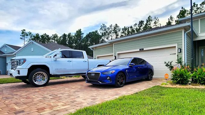 A white GMC pickup truck and a blue Hyundai Genesis sedan parked on a brick driveway in front of a house.