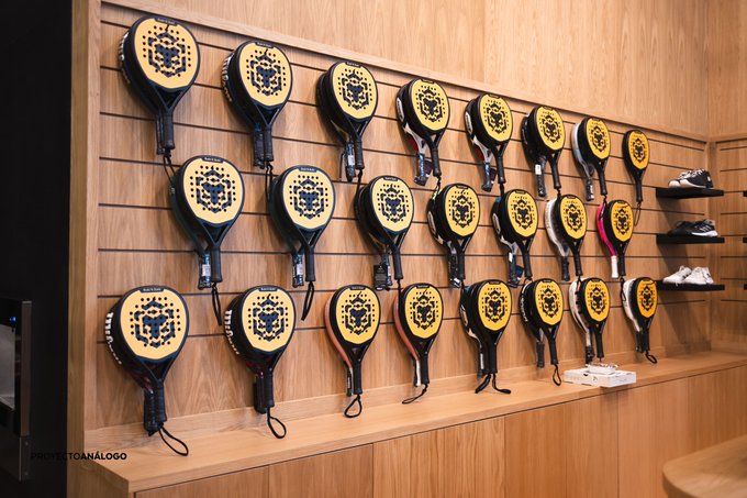 A wooden display wall in a store features rows of black and yellow padel rackets hung in an organized grid, with a few pairs of shoes on shelves to the right. A watermark in the bottom left reads "PHOTOS: @CANALPADEL" or similar, fitting the "Padel N Padel" theme by showcasing a retail selection of padel equipment.