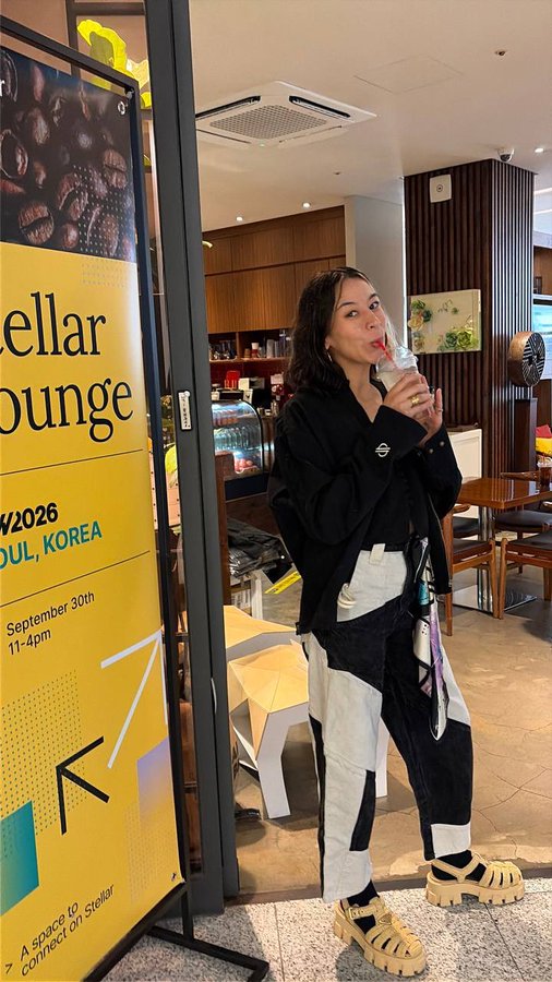 A woman wearing a black jacket and black-and-white pants drinks from a bottle while standing in what appears to be a 'Stellar Lounge' (mentions KW2026 in Seoul, Korea), with a yellow banner visible to her left.