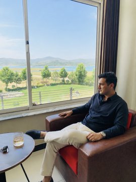 The image shows a man sitting comfortably in an armchair with his legs crossed while looking out of a window at a beautiful view of mountains. The man appears to be enjoying the scenery, possibly contemplating or taking a break from work. On the table next to him, there is a glass and a cell phone, suggesting that he might have been using it for communication or capturing the moment with his camera.