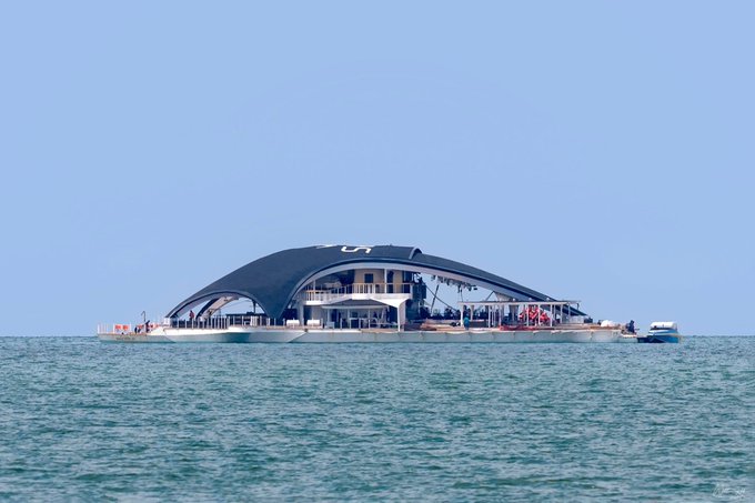 A large, modern floating structure with a curved black roof sits on the open ocean under a clear sky, with small boats nearby.