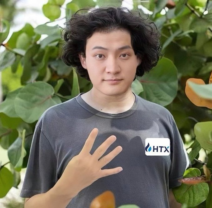 A young man with curly dark hair wearing a gray t-shirt with an "HTX" logo holds up four fingers against a backdrop of green leaves.