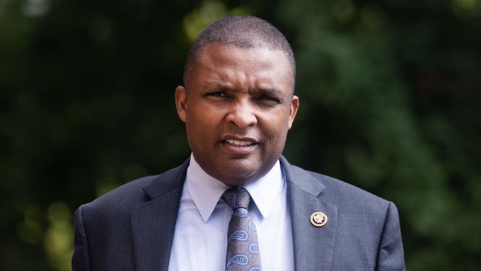 Portrait of a man in a grey suit and patterned tie wearing a gold lapel pin, photographed outdoors against a blurred green background.