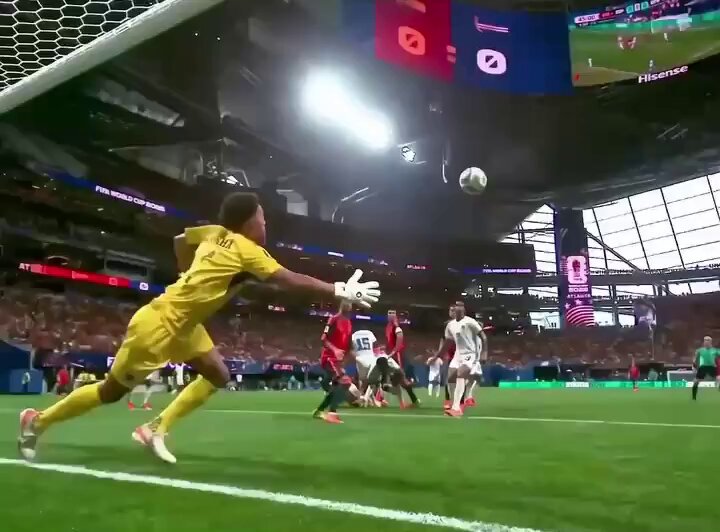 The image shows a soccer game taking place with players from two different teams engaged in an intense match. One player is wearing yellow and appears to be diving towards the ball while another player in white is also diving for the ball, both of them trying to gain control over it. The goalkeeper is seen holding his hands up, attempting to block the ball from entering the goal. In the background, there are two TVs displaying the game's progress, with one showing a close shot of the ball and the other showing the players' positions on the field.