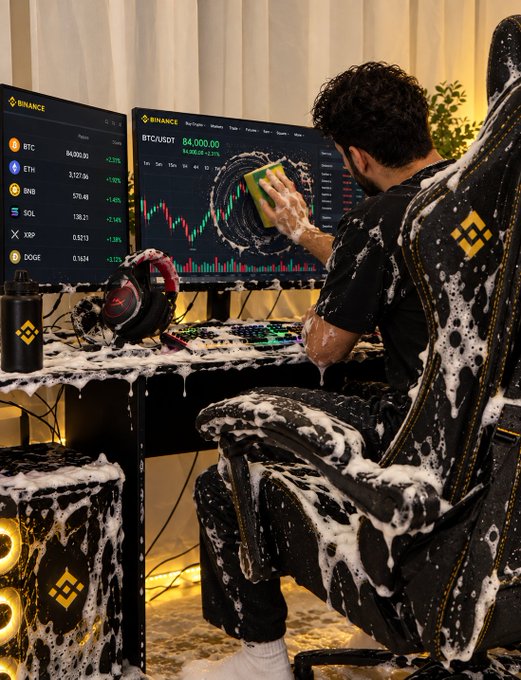 A trader covered in foam scrubs a screen displaying Bitcoin at $84,000 and the BTC/USDT trading chart, alongside Binance interfaces listing tickers like BTC, ETH, BNB, SOL, XRP, and DOGE—visually interpreting "clean up your old trades" as a literal sudsy wash of his cryptocurrency workspace.