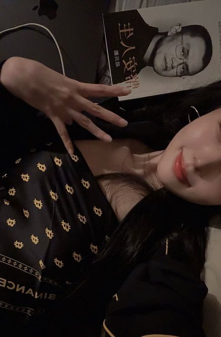 A person poses with a Chinese edition of "The Book of CZ" while wearing a Binance-branded top, illustrating the book's giveaway promotion. The image ties to the post's mention of CZ as the "on-chain wish-granting god" and the $FOMA project's animated adaptation and giveaway campaign for the book.