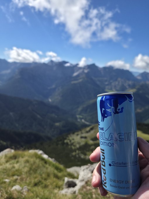 A hand holding a light blue Red Bull 'Glacier Edition' energy drink can in the foreground, with a stunning mountain landscape and partly cloudy sky in the background.