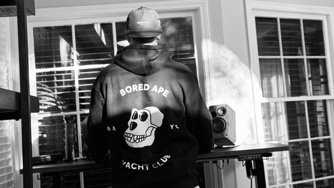 A black-and-white photo of a person wearing a "BORED APE YACHT CLUB" hoodie and a cap, standing with their back to the camera facing a window and a studio monitor.