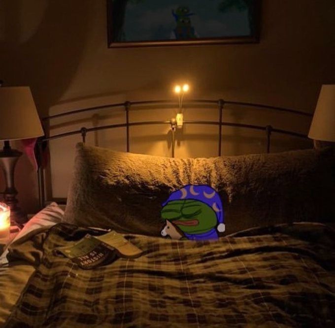 A dimly lit bedroom with a metal-framed bed, plaid bedding, two lamps, and a Pepe the Frog plush toy resting against the pillows.