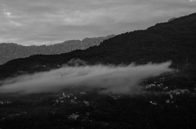 A black-and-white photograph shows mist or low clouds draped over dark mountains with a small village nestled below.