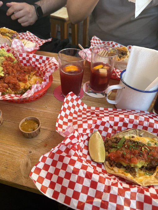 A table set with red and white checkered paper holding plates of tacos and burritos, with two glasses of a dark drink garnished with orange slices.