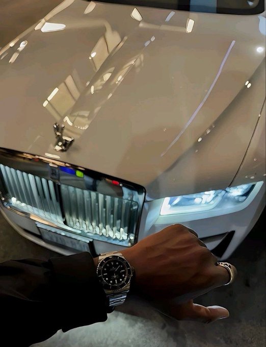 A close-up of a person's wrist wearing a luxury silver-toned watch, posed against the illuminated front grille and hood of a luxury car, likely a Rolls-Royce, in a showroom setting.