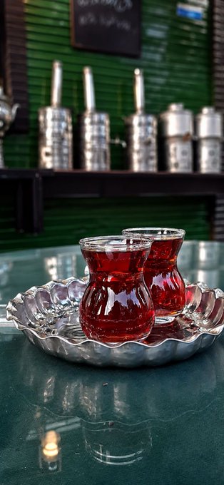 Two traditional Turkish tea glasses filled with red tea sit on a silver tray on a green table, with metallic tea dispensers visible in the background.