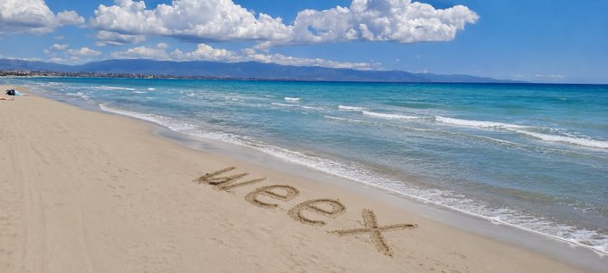 A beach scene with the word 'Meex' written in the sand near the shoreline, with turquoise water and mountains in the background.