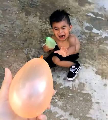 A young shirtless boy cries and reaches out in fear as a hand holds an orange balloon toward him on a concrete surface.