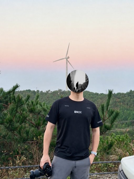 A person wearing a black OKX t-shirt stands outdoors holding a camera, with their face obscured by a circular blur, against a backdrop of pine trees and a wind turbine at sunset.