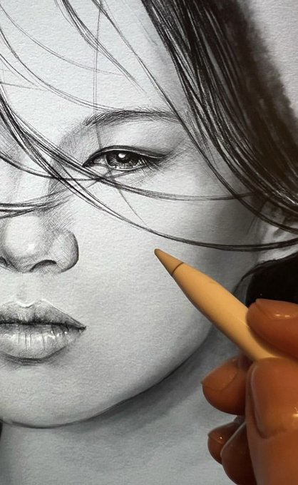 A close-up photograph of a detailed pencil drawing of a woman's face, with a hand holding a blending stump working on the cheek area.