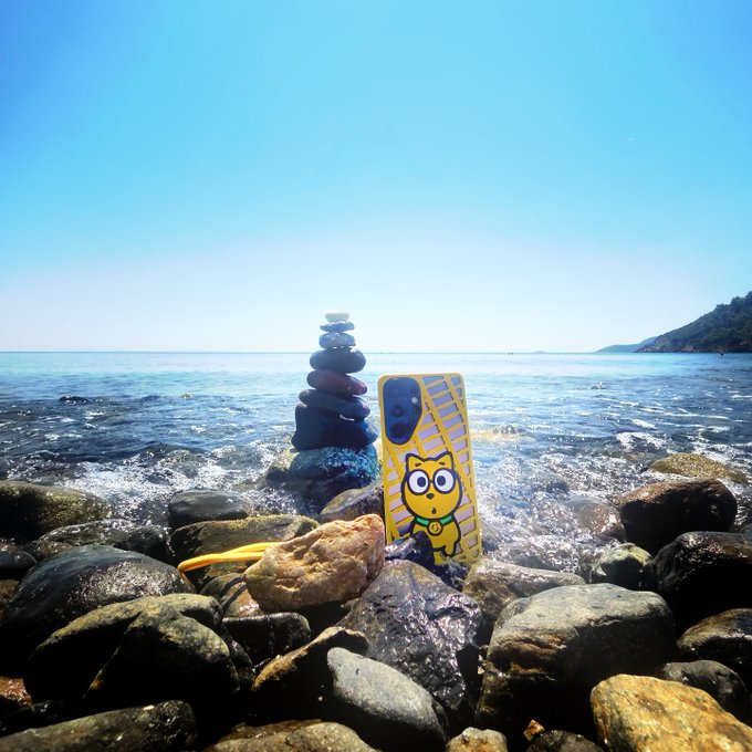 A photo of a phone case with a yellow cat design standing among rocks at a beach, next to a small stack of balanced stones with the sea in the background.