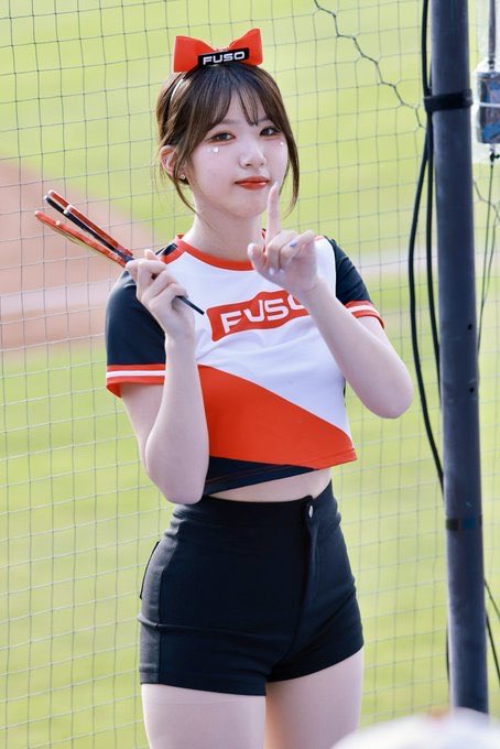 A young woman wearing a FUSO cheerleading uniform and bow, posing with her finger to her lips in a 'shh' gesture while holding pom-poms at a baseball stadium.